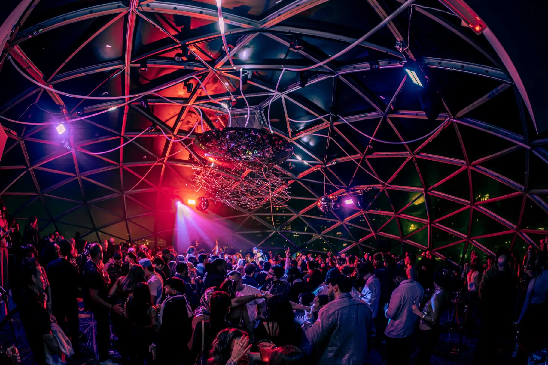 Packed dance floor at a Reunion event, lit in red and violet with the Minneapolis skyline visible through the glass